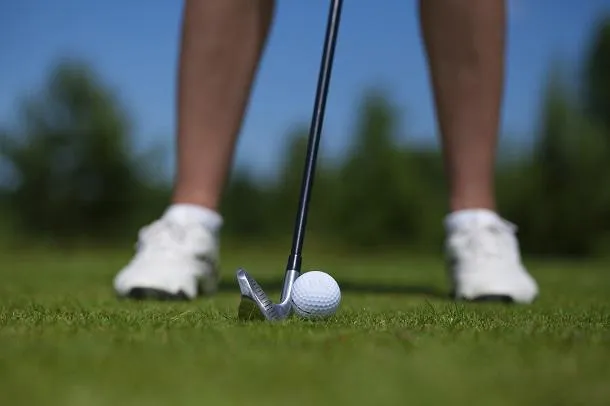 un gros plan d'une balle de golf contre l'herbe verte vibrante, avec le fer d'un golfeur et ses jambes