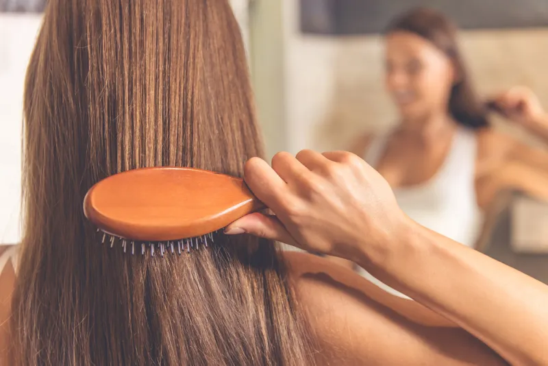 back view of beautiful young woman in white undershirt combing her hair and smiling while looking into the mirror in bathroom