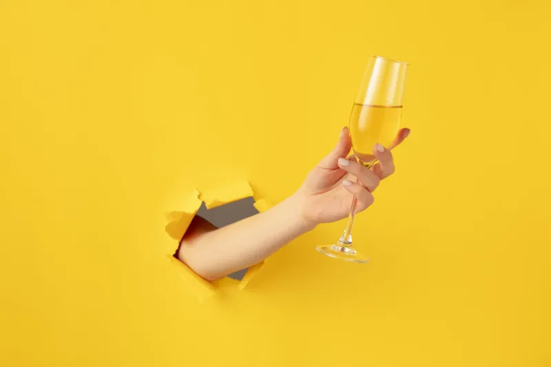 beautiful woman girl hand with champagne flute glass breaks through yellow paper background copy space for ad, design, text concept of holidays, celebrations and greeting