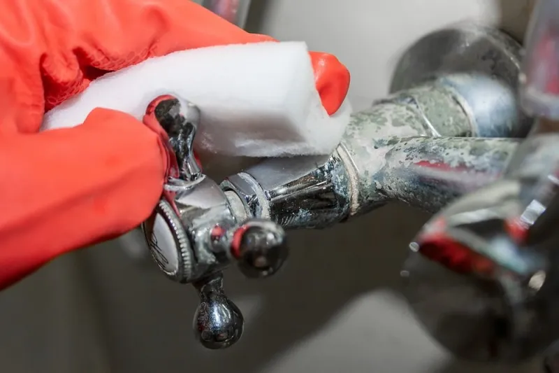 a red-gloved hand rubs a hot water knob on a chrome-plated faucet covered with limestone with a white melamine sponge selective focus closeup view
