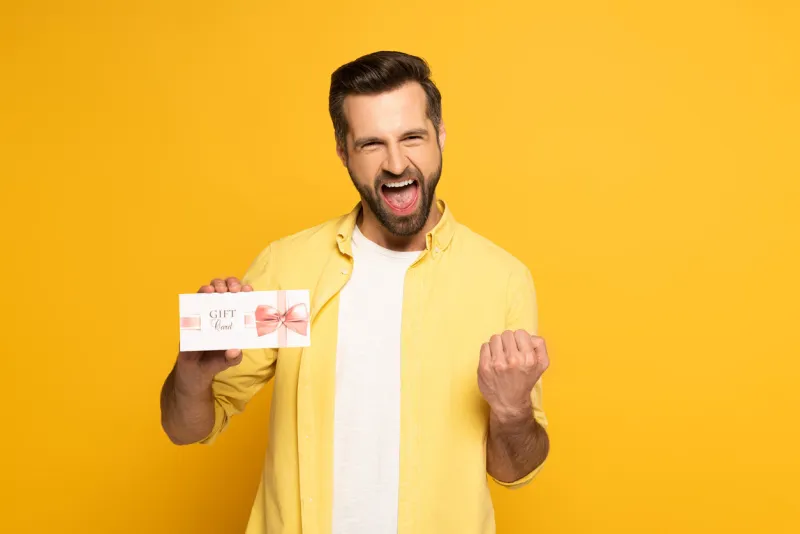 cheerful man showing yeah gesture and holding gift card on yellow background