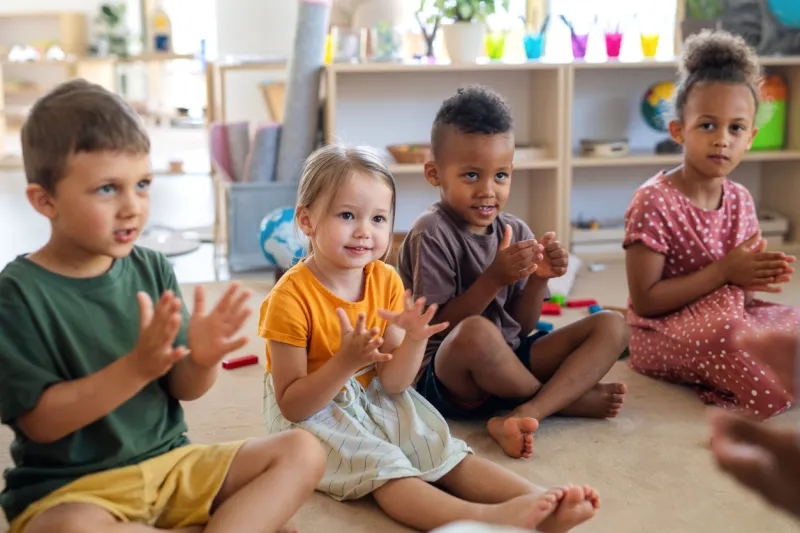 a group of small nursery school children sitting on floor indoors in classroom, clapping
