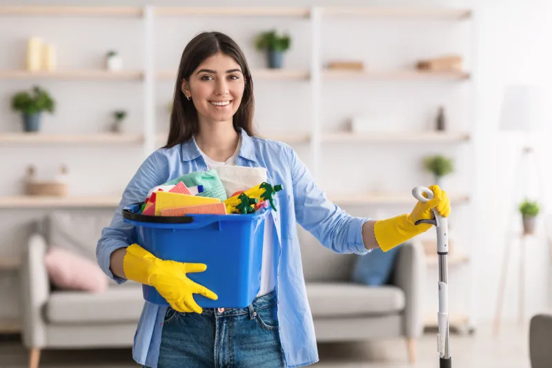 portrait of smiling millennial lady holding bucket with cleaning supplies and mop, posing and looking at camera standing in living room professional cleaning service specialist wearing rubber gloves