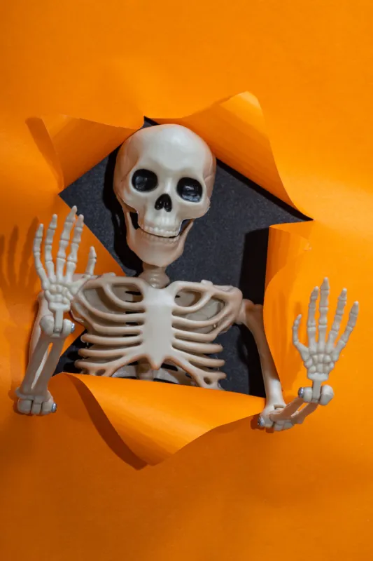 happy halloween cute little toy skeleton waving bony hands through a torn paper hole on a bright orange background, vertical frame, minimalism hello halloween