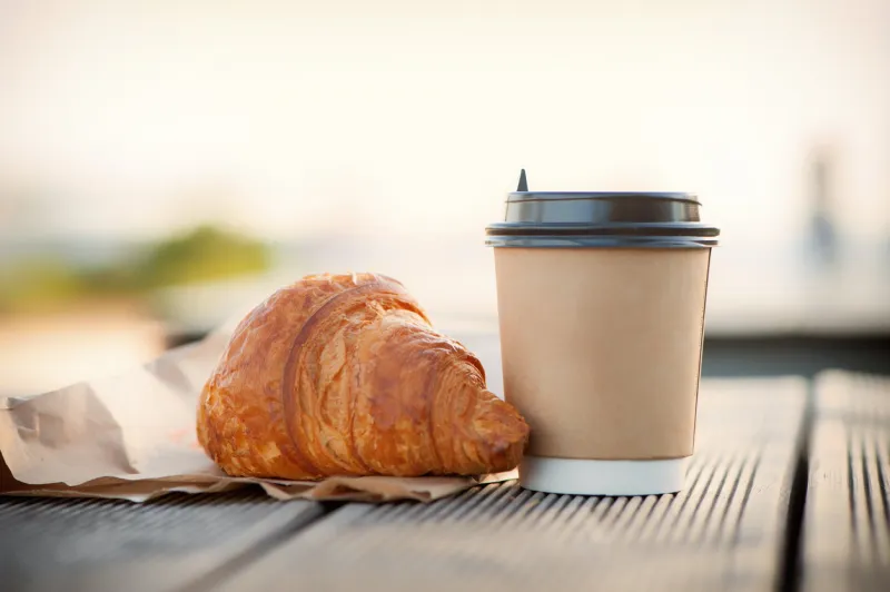 eco paper cup with coffee or tea and croissant on kraft paper, on a wooden table breakfast on the street, in the park, on a bench