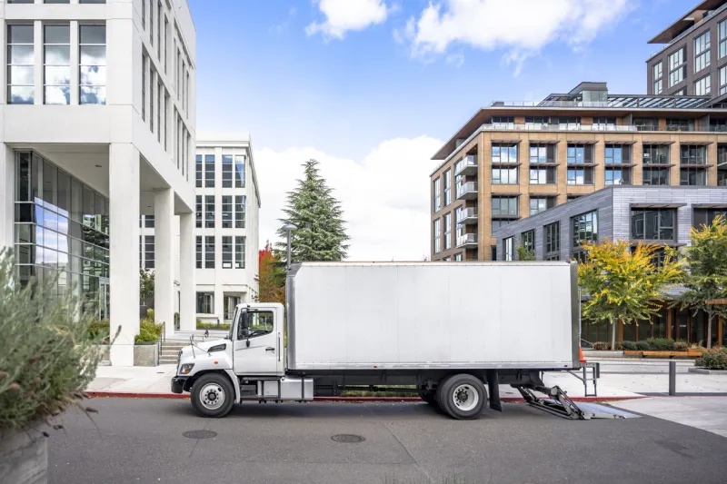 white middle class relocation rig day cab semi truck tractor with long spacious box trailer standing on the urban city street with multilevel apartment and office buildings unloading delivered goods