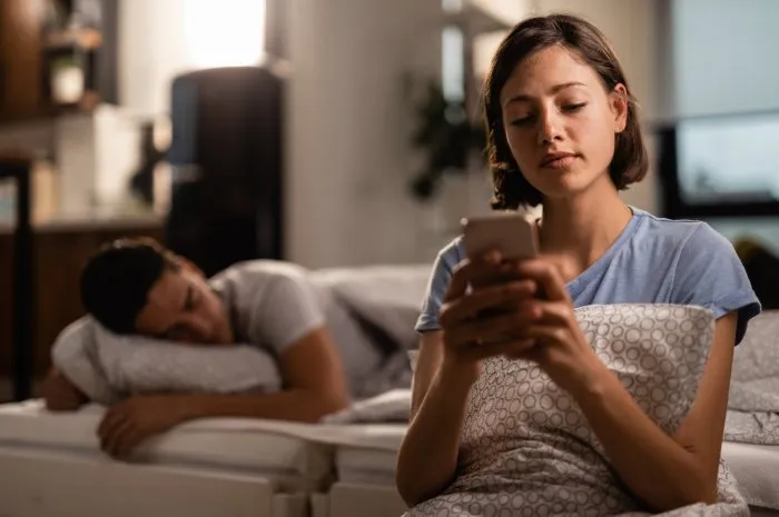 young woman text messaging on cell phone while her boyfriend is sleeping on the bed behind her