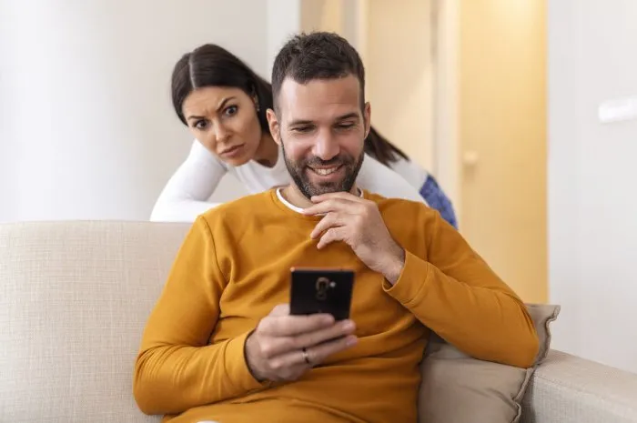 cheater man dating on line with a smart phone and girlfriend is spying sitting on a sofa at home