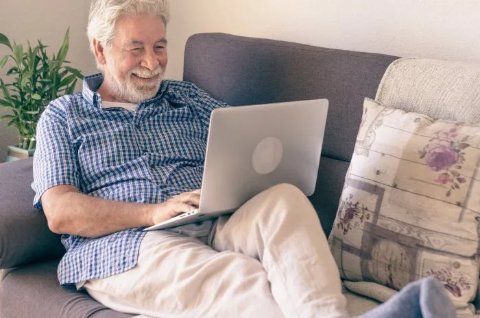 smiling adult mature senior man with beard resting on sofa at home using laptop relaxed attractive elderly grandfather enjoying free time and technology