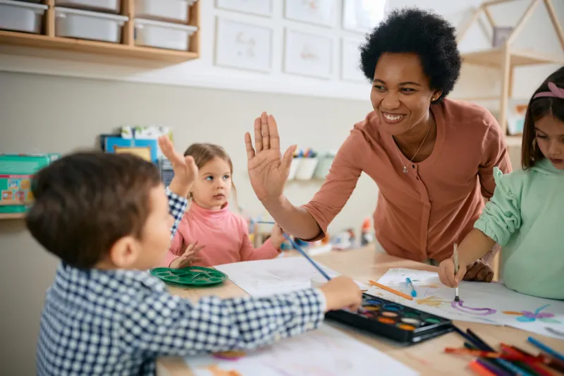 happy african american teacher and small boy giving high-five during art class at kindergarten