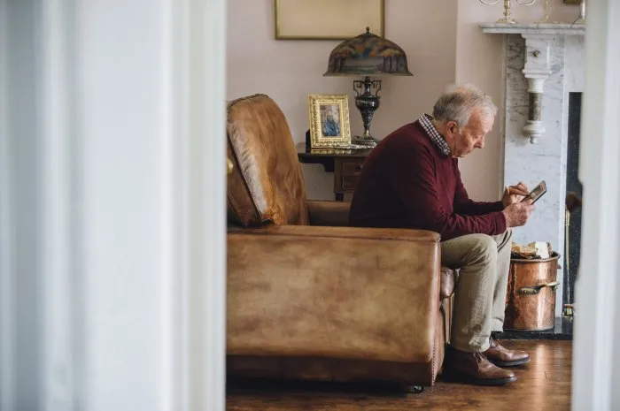 senior man is sitting in an armchair in the living room of his home, holding and looking at an old photo with a sad expression