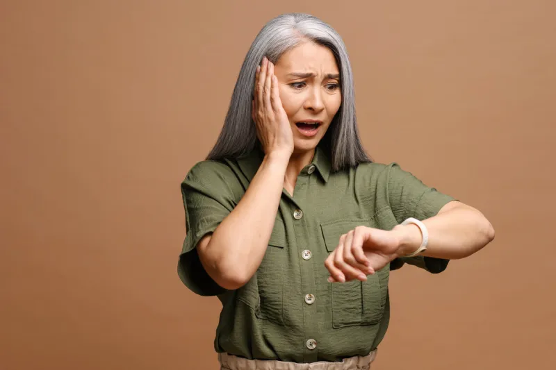 portrait of shocked mature woman holding hand with wrist watch and looking at it with surprised expression isolated on a brown background