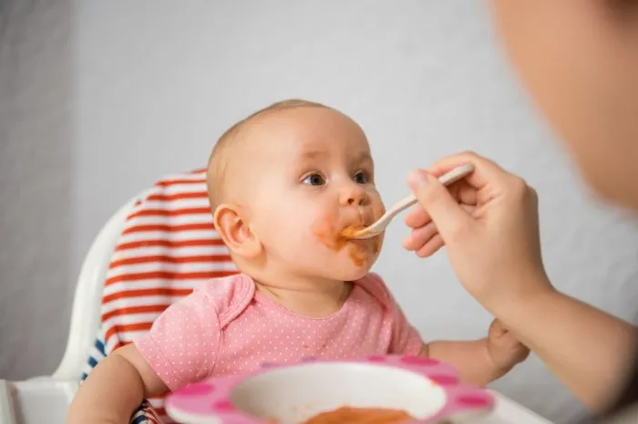 mother feeding her baby girl with spoon in high chair, they enjoy in togetherness
