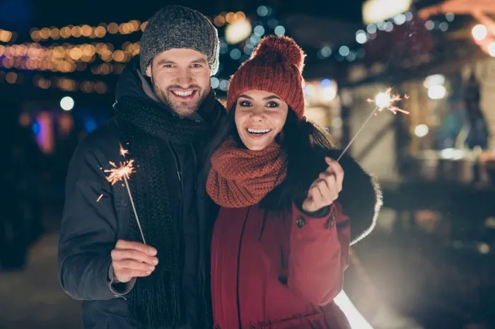 photo of funny couple at x-mas celebration in park holding magic sparklers, excited to meet newyear midnight wearing warm coats knitted caps and scarfs outdoors