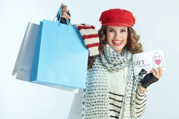 smiling young female with long brunette hair in sweater, scarf and red hat showing shopping bags with sweaters and discount coupon against winter light blue background