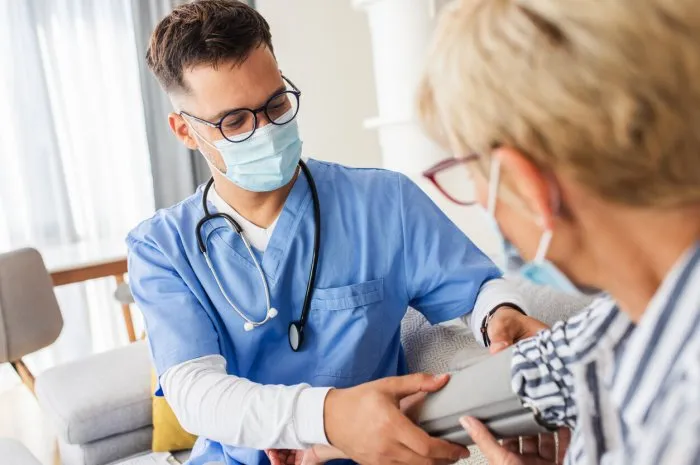 male nurse measures blood pressure to senior woman with mask while being in a home visit