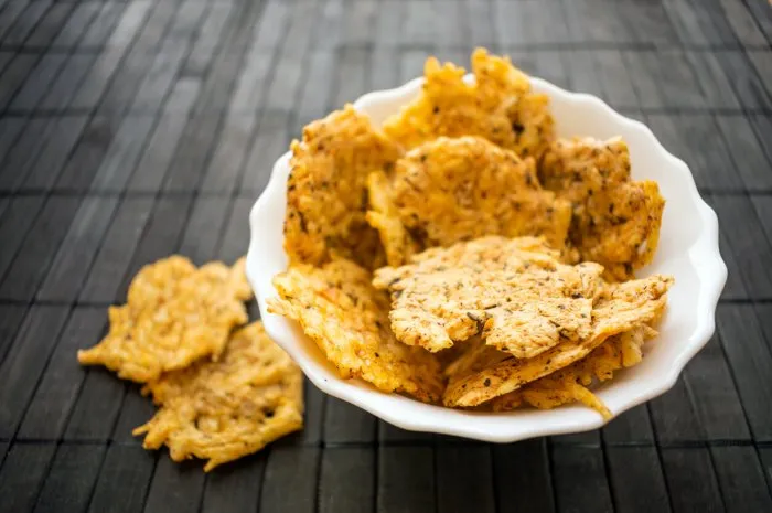 cheese chips snack in a round white bowl on a black wooden background grain free dippable crispy cheddar cheese chips, keto & low carb crunchy chip to snack from cheddar, parmesan, asiago, herbs, spices