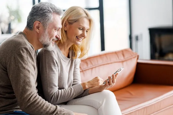 serene middle-aged spouses using smartphone sitting on sofa in living room, grey-haired husband and beautiful wife making order online,romantic mature couple using mobile app for booking vacation tour