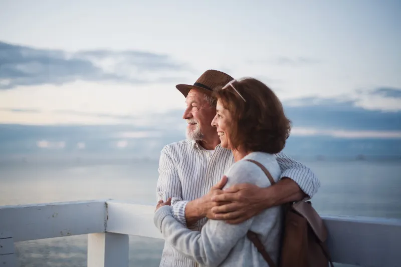 portrait of a happy senior couple in love hugging outdoors on pier by sea, looking at view, summer holiday