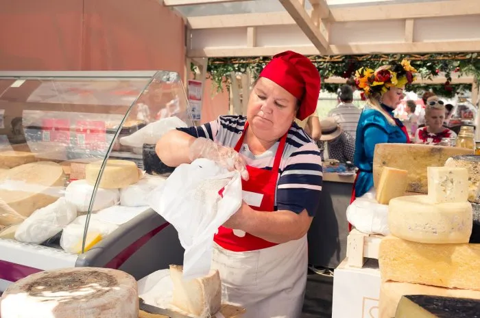 moscow, russia, october 01, 2022  a middle-aged female salesman wraps a piece of cheese in paper while standing behind a store counter at a grocery market a sunny summer day