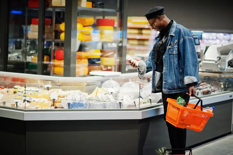 stylish casual african american man at jeans jacket and black beret holding basket, standing near cheese fridge and shopping at supermarket