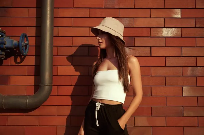 confident young female wearing trendy bucket hat standing with hand in pocket against wall of building in city and looking away
