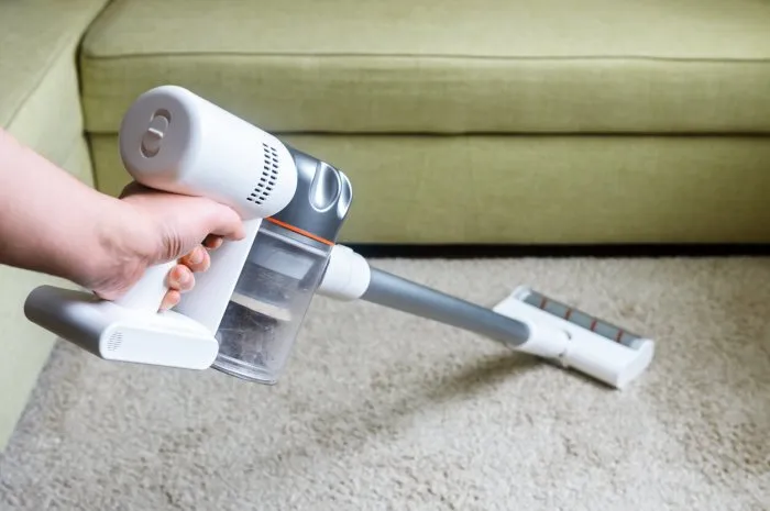 wireless vacuum cleaner used on carpet in room housework with new upright hoover person holds modern white vacuum cleaner by sofa home cleaning, care and technology concept