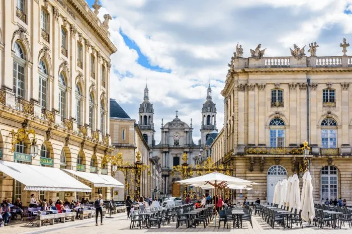 nancy, france - september 12, 2019  sunshine on the terrace of the sidewalk cafe of the grand hotel on the place stanislas closed by a gilded wrought iron gate with nancy cathedral in the background