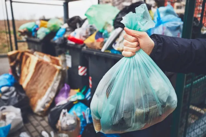 hand with garbage against full trash cans with rubbish bags overflowing onto the pavement