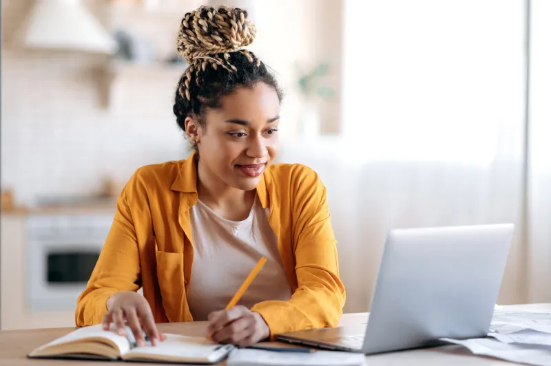 focused cute stylish african american female student with afro dreadlocks, studying remotely from home, using a laptop, taking notes on notepad during online lesson, e-learning concept, smiling