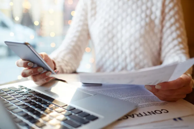 closeup of unrecognizable busy woman multitasking  holding documents in hand, typing message in smartphone and reviewing contracts and statistics while working with laptop at cafe table in evening