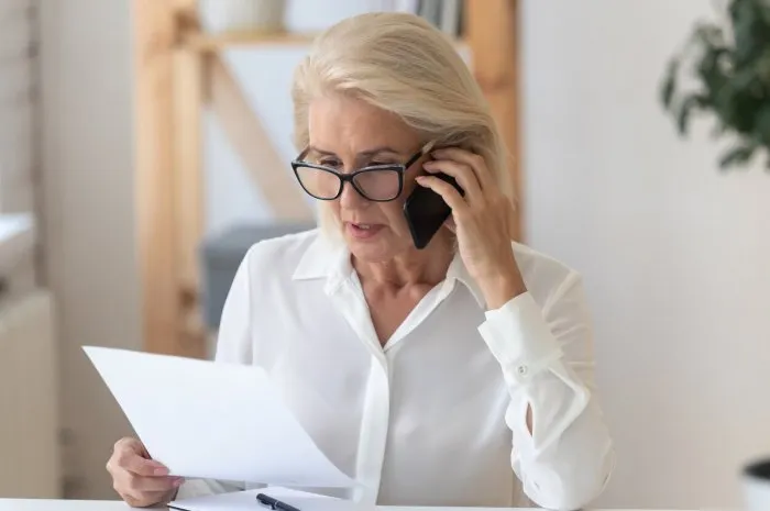 concentrated senior businesswoman in glasses sit at office desk talk on cellphone reading paperwork document, focused middle-aged woman speak with client on smartphone discuss contract