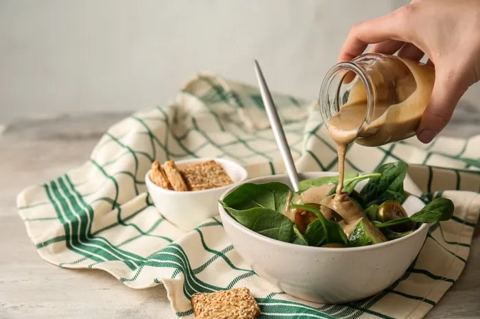 pouring of tasty tahini from jar onto fresh vegetables in bowl
