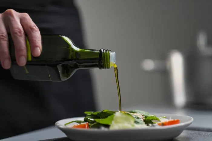 a female chef in a white uniform and a black apron in the restaurant kitchen cooking the cook pours olive oil from a green bottle grey background