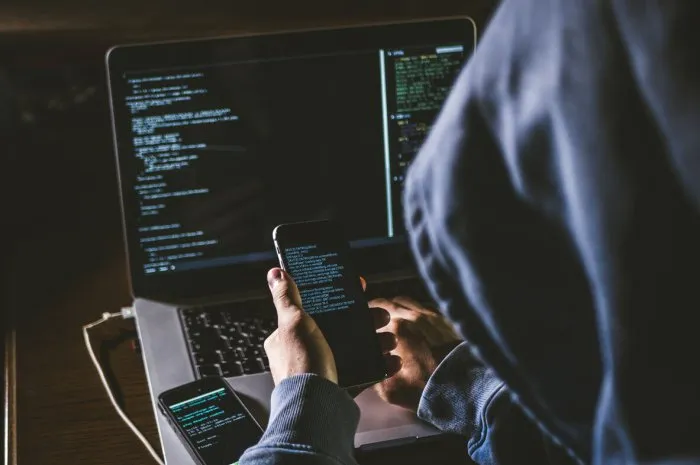 anonymous person in the hood sitting in front of computer working with laptop and mobile phone