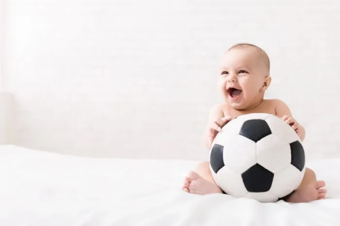 little football lover adorable baby sitting on bed with big soccer ball and laughing, copy space