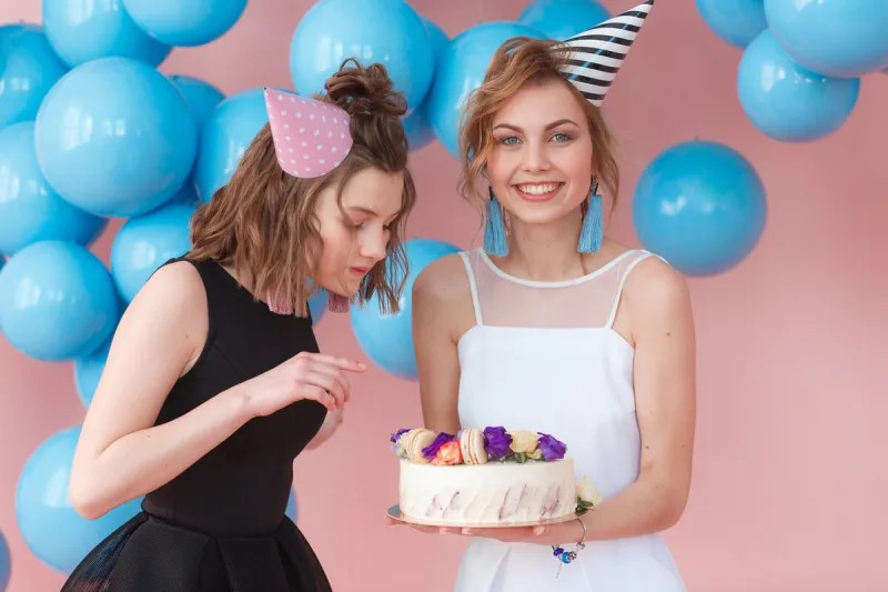 two teenage girls in party hat holding cake isolated on pink background and blue balloons