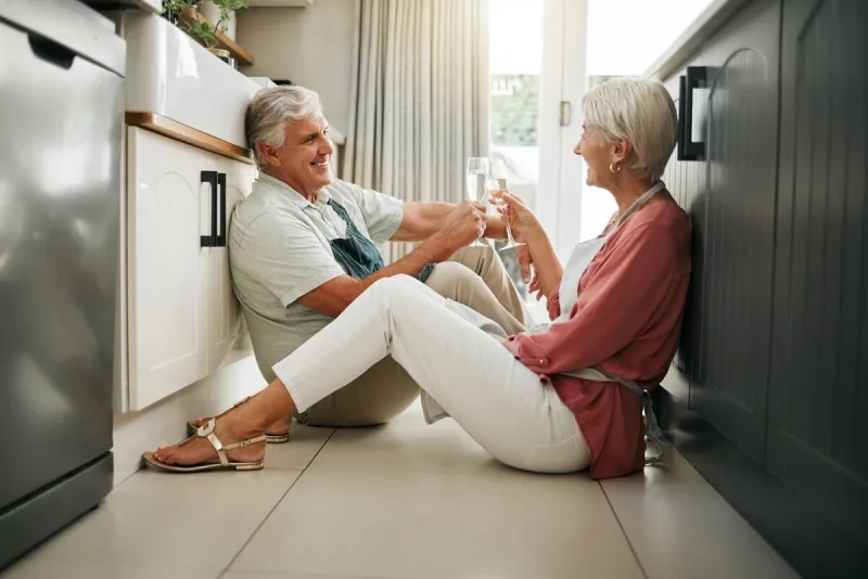 champagne celebration, toast and senior couple having a drink to celebrate retirement on the floor of kitchen in home elderly man and woman happy on anniversary of marriage with cheers and alcohol