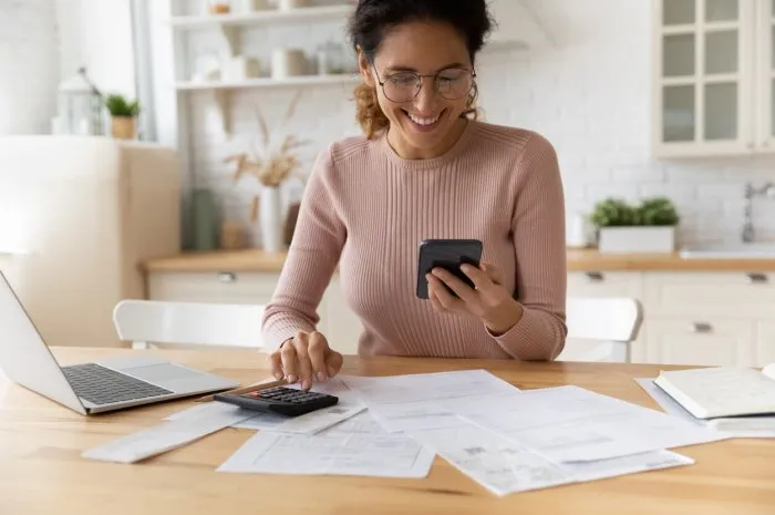 smiling young 30s woman in eyewear looking at smartphone screen, feeling satisfied with fast secure online service, paying household bills taxes or insurance, managing budget, calculating expenses