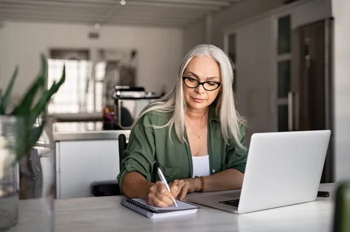 senior stylish woman taking notes in notebook while using laptop at home old freelancer writing details on book while working on laptop in living room focused cool lady writing notary in notepad
