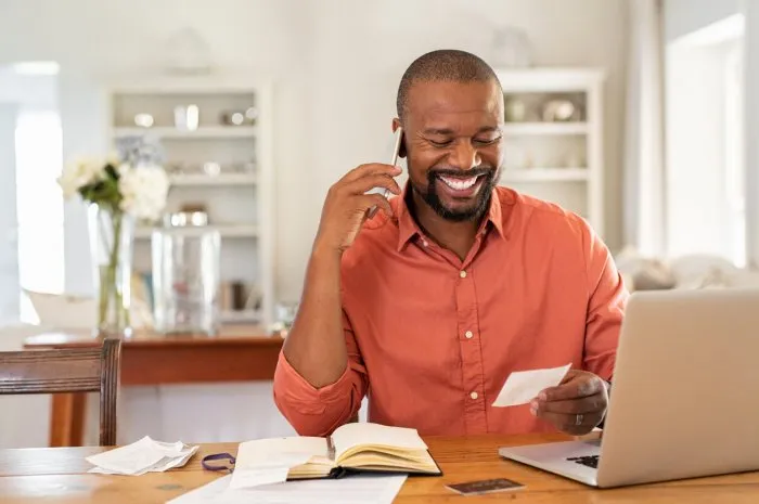 smiling african man using laptop at home while checking home finance happy mature man looking at invoice while talking on phone with bank man checking receipt and bill while talking at phone with insurance agentr