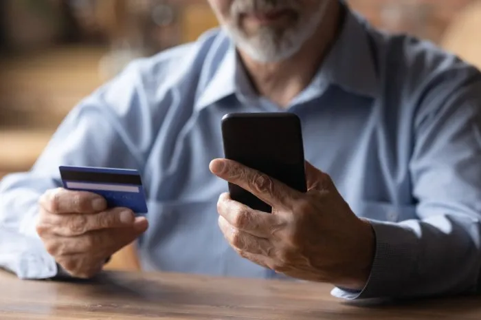 close up mature man using phone, holding plastic credit or debit card, senior grey haired customer making secure internet payment, shopping or browsing online banking service, entering information