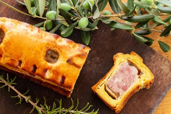 top view of a pate en croute or pâté en croûte or meat pie, with rosemary twig and green olives on branch with leaves over a dark wooden cutting board and a used oak wood background french traditional appetiser