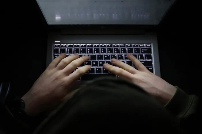 a simple, very dark night time image of hands on an illuminated keyboard typing shady person wearing a hood at a computer or laptop in the dark