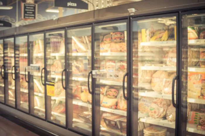 blurred variety pack of processed pizza at retail store in america frozen food section in huge glass door aisle freezer full assortment of frozen pizza in local supermarket, defocused background