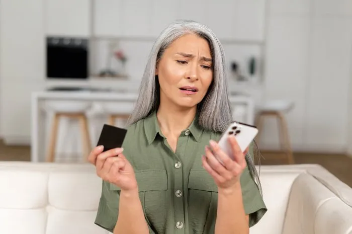 frustrated unhappy gray-haired mature woman sitting on sofa, holding smartphone and credit card checking card balance, senior female has not enough money for online transaction, financial problem