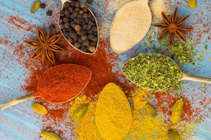 milled spices - garlic, turmeric, paprika, anise, oregano, cardamom round of golden spoons on blue wooden table top view, close-up