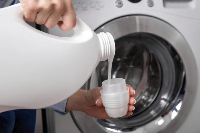 against background of drum of steel-colored washing machine, woman pours liquid washing gel into plastic cap a girl in a white t-shirt carefully pours a transparent conditioner for flattening laundry