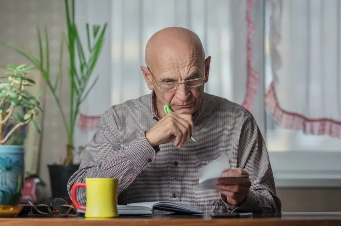 pensive older man wearing glasses checking financial document sitting at kitchen table at home