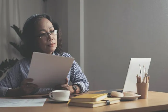 seriously senior woman tworking at home, looking at laptop and waving paper correspondence, sitting at home, free space